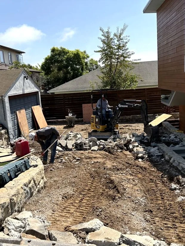 A mini excavator breaking out an old driveway beside a stone retaining wall
